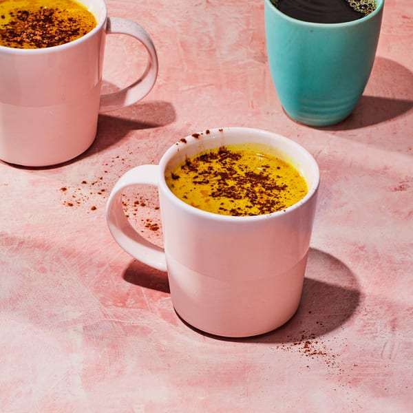 Three mugs, two pink with golden milk and one teal with coffee, on a pink textured surface.