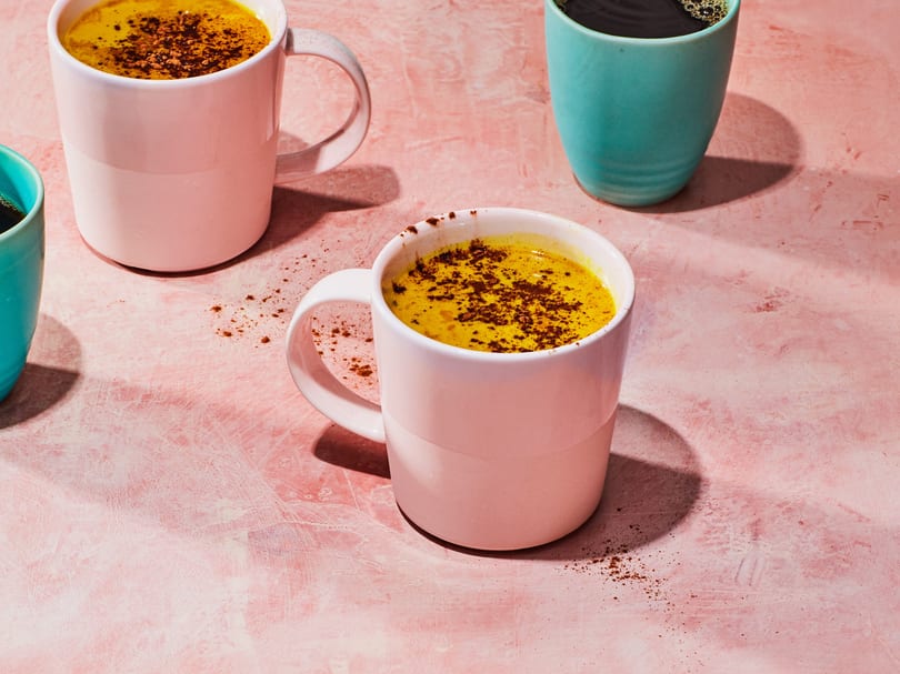 Three mugs, two pink with golden milk and one teal with coffee, on a pink textured surface.