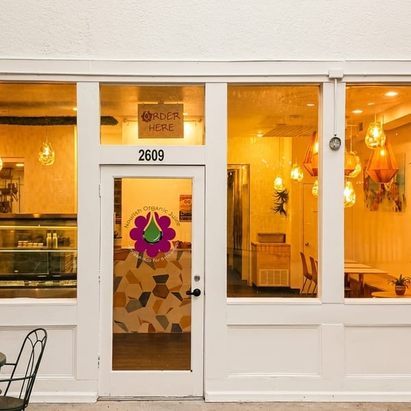 Exterior of Nourish Organic Juice, a white storefront with large windows, outdoor seating, and warm interior lighting.