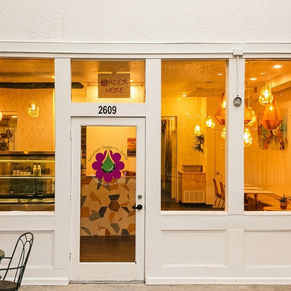 Exterior of Nourish Organic Juice, a white storefront with large windows, outdoor seating, and warm interior lighting.