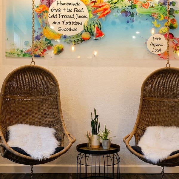 Two wicker hanging chairs with fluffy white cushions flank a small table with plants, under a fruit-themed sign.