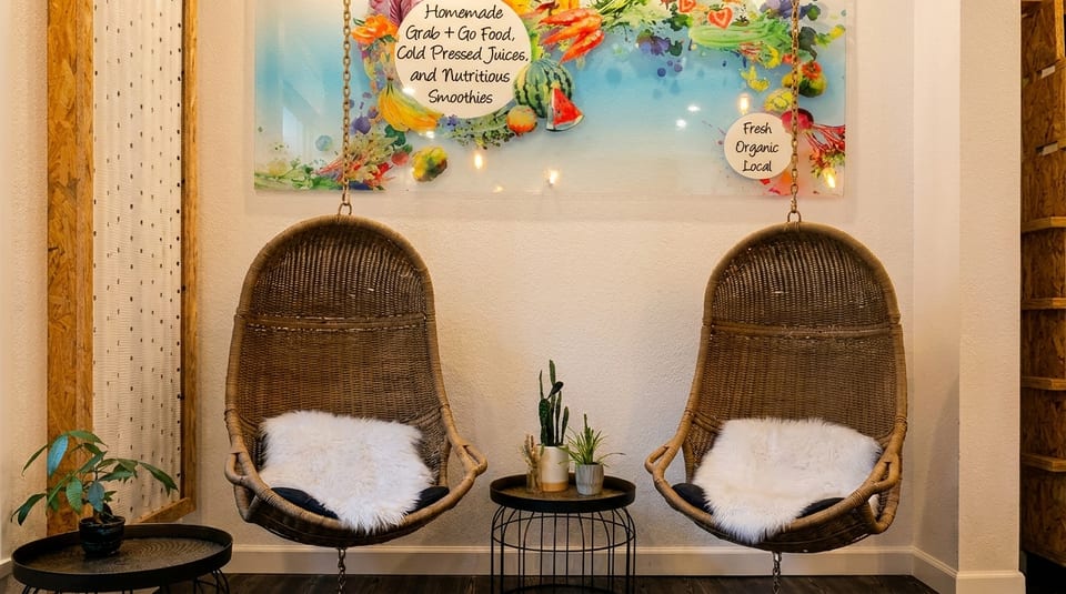 Two wicker hanging chairs with white furry cushions flank a small table with plants, under a fruit-themed sign.