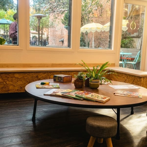Cozy restaurant interior with a low oval table, small stools, plants, and natural light from large windows.