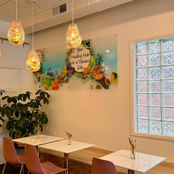 A brightly lit cafe interior with colorful pendant lights, white tables, and a lush green plant wall.