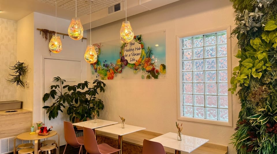 A bright cafe interior with colorful pendant lights, green plants, and white tables with pink chairs.
