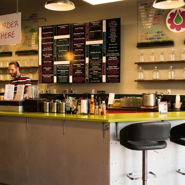 A man behind a bright green counter with bar stools, menu boards, and shelves of bottles.