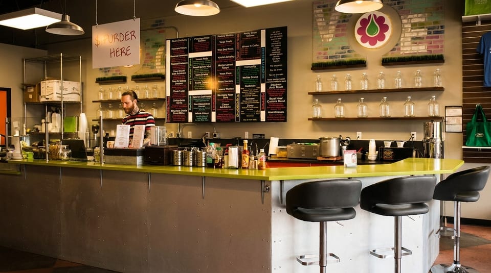 A man behind a bright green counter with bar stools, menu boards, and shelves of bottles.