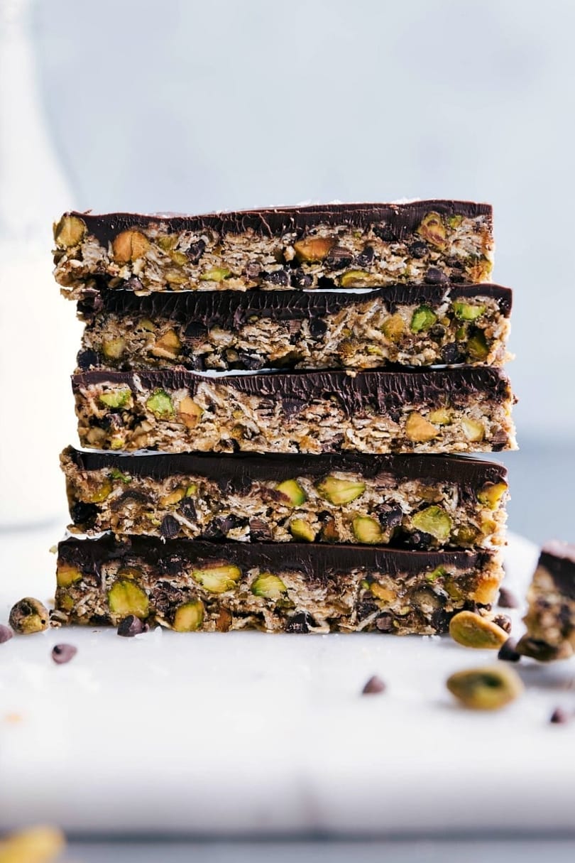 Stack of five chocolate-topped granola bars with pistachios and almonds on a white surface.
