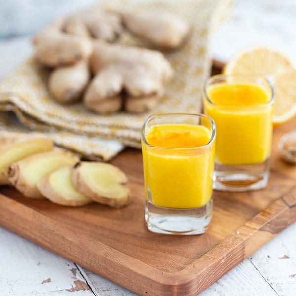Two ginger shots on a wooden tray with sliced ginger, whole ginger, and lemon.