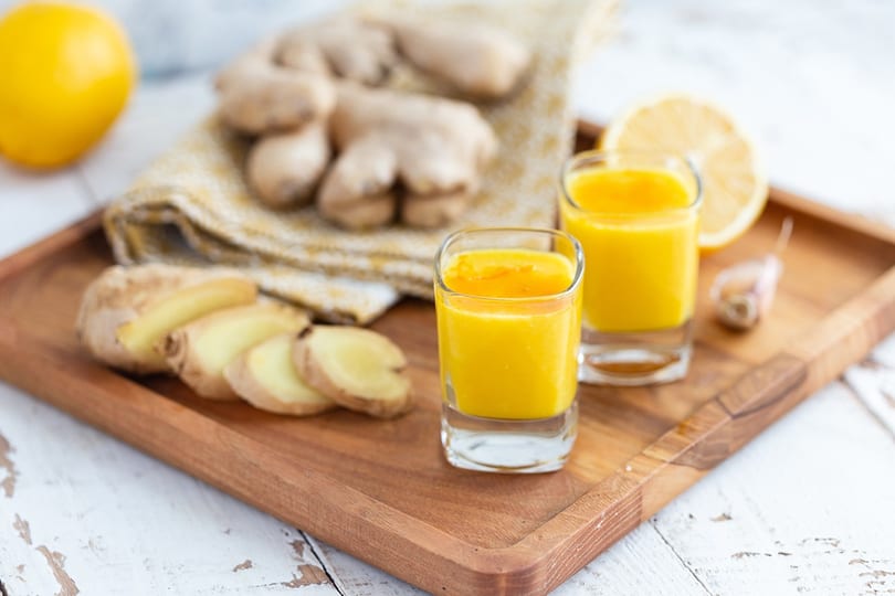 Two ginger shots on a wooden tray with sliced ginger, whole ginger, and lemon.